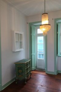 Das Wohnzimmer des Apartamento Lino im 2. Stock. Der Blick geht in Richtung Wohnzimmer und zeigt eine Balkontür, ein Fenster, die Decke mit Original erhaltenem Stuck der einen Engel darstellt. Die Wände sind weiß, der antike, restaurierte Dielenboden in warmen braunton mit geölter Oberfläche. Ein rotes Schlafsofa steht rechts, links eine antike Kommode die modern in hellem grün und gold hübsch restauriert ist. An der Decke ist ein antiker Kronleuchter der das Wohnambiente angenehm unterstreicht. Der Raum ist sehr hübsch, hell und frisch.
