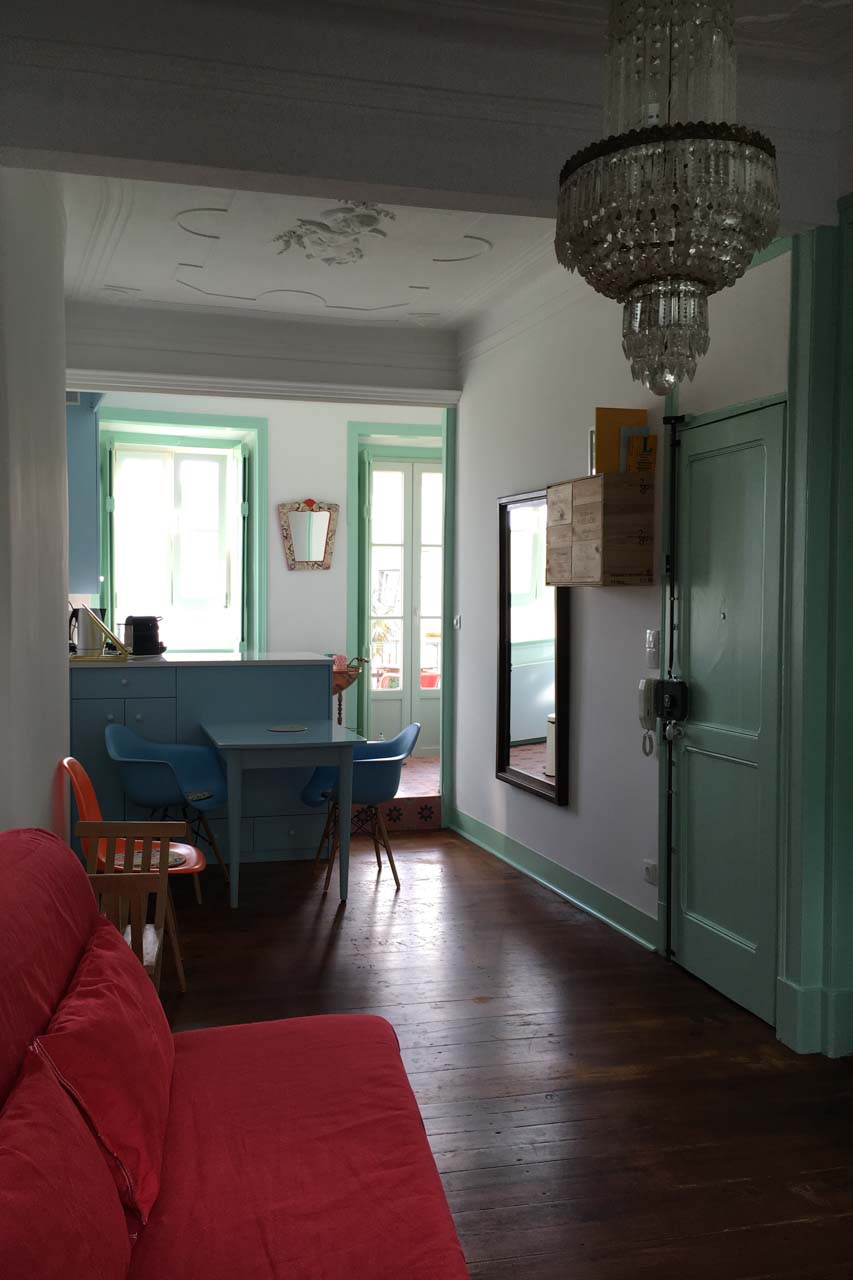 Das Wohnzimmer des Apartamento Lino im 2. Stock. Der Blick geht in Richtung Küche und Bad, er zeigt die Balkontür die zum Balkon auf der Rückseite des Hauses führt, das Küchenfenster, eine Stufe die das Wohnzimmer von der Küche trennt,die Decke mit Original erhaltenem Stuck der einen Engel darstellt. Die Wände sind weiß, alle Türen und Fensterrahmen in zartem lindgrün, die Küchenmöbel passend in pastellfarbenem hellblau, der antike, restaurierte Dielenboden in warmen braunton mit geölter Oberfläche. Ein rotes Schlafsofa steht links, rechts eine antike Kommode die modern in hellem grün und gold hübsch restauriert ist. An der Decke ist ein antiker Kronleuchter der das Wohnambiente angenehm unterstreicht. Der Raum ist sehr hübsch, hell und frisch.