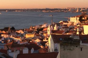 Lissabon, sein historisches Herz die Alfama, der Tejo ist in Abendlicht getaucht. Der Fluß Tejo liegt ruhig und man sieht im Vordergrund die Dächer der Altstadt und seine weißen Kirchengebäude. Ein Kran der an einer Baustelle aufgebaut ist die am Ufer des Tejo liegt ragt aus der Höhe.