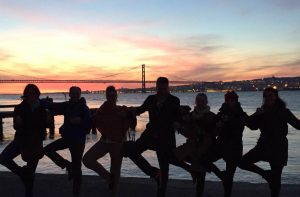 Yoga Städtereise nach Lissabon, hier ist die Gruppe im Abendlicht am Tejo in einer Reihe aufgestellt und alle stehen auf einem Bein. Das zweite Bein ist angewinkelt, Fußsohle ans Standbein geheftet. Im Hintergrund ist die Brücke 25. April, der gelb-orange Abendhimmel und der Tejo. Die Gruppe ist nur in Umrissen zu sehen.
