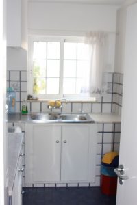 Die Küche im Casa Valéria sieht man auf diesem Bild. Links ist eine Küchenzeile in weiß mit einem Kühlschrank, Unter-und Oberschränken, daran schliesst der Herd mit Backofen und eine Spüle an. Die Küche hat ein Fenster und Tageslicht.